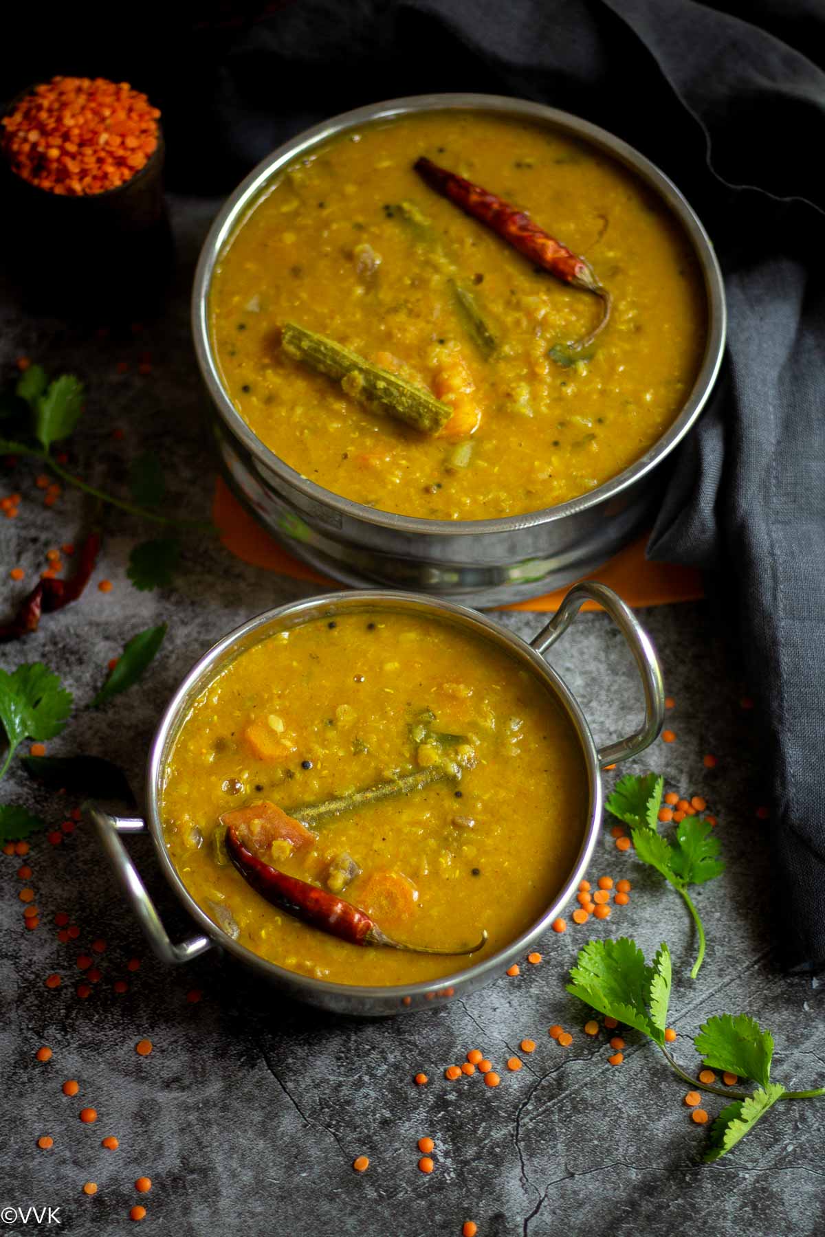 overhead shot of masoor dal sambar served in two vessels with gray fabric on one side and masoor dal in measuring cup on the other side