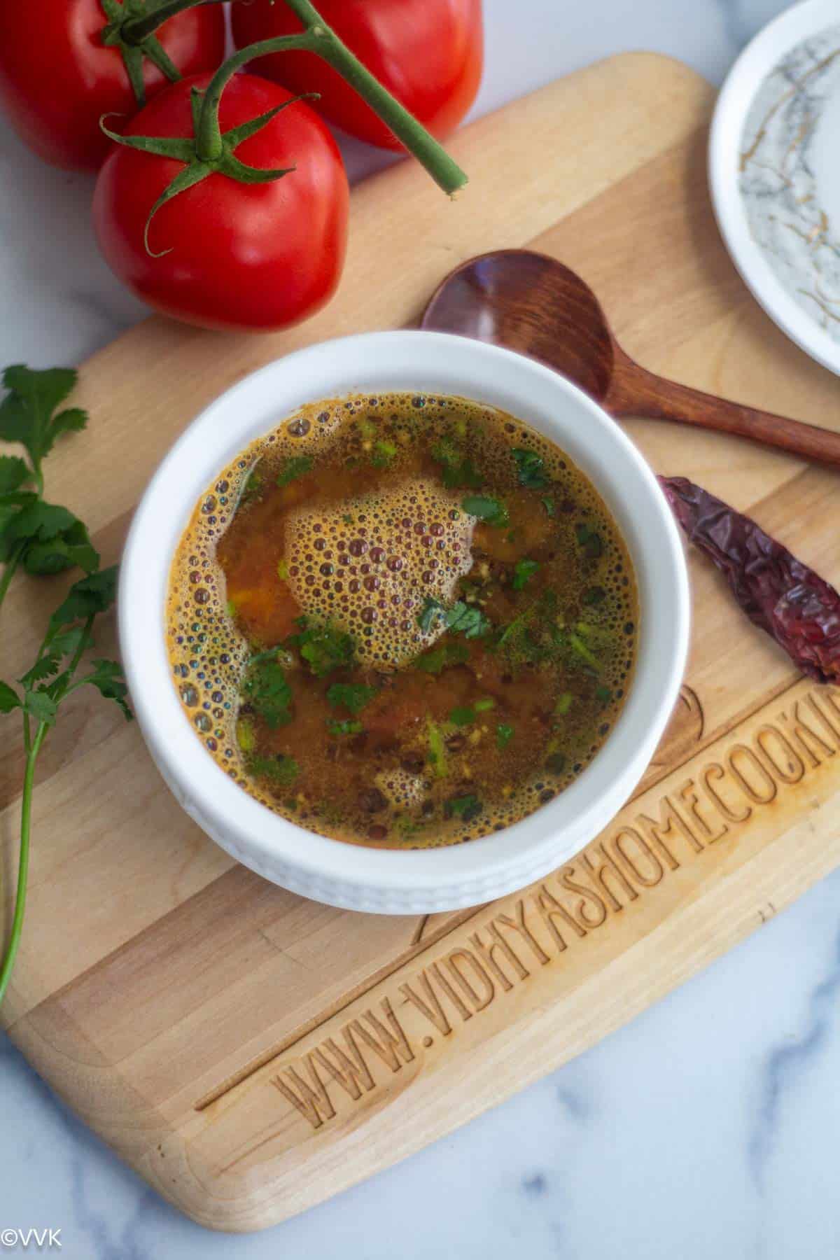 simple mysuru rasam served in white bowl placed on wooden board with tomatoes on the side