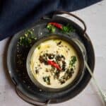 square image of urad dal raita served in black ceramic ware placed on iron tray with chilies and cilantro scattered