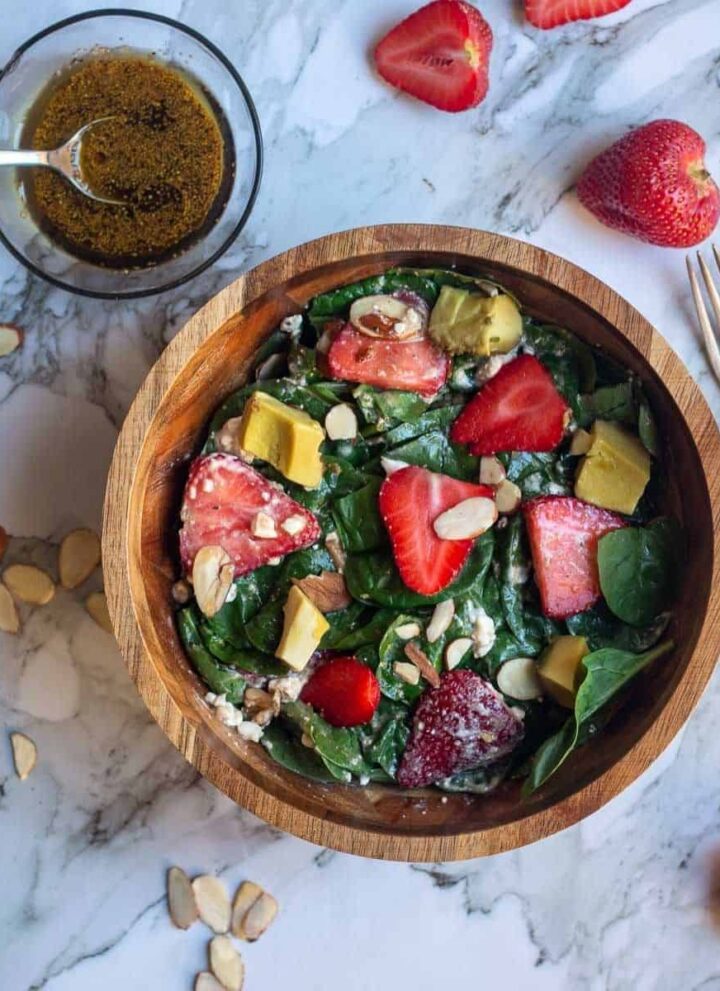square image of spinach and strawberry salad in a wooden bowl with the dressing on the side