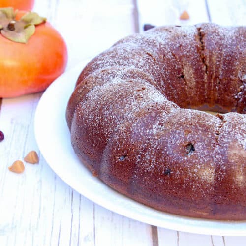 Eggless Persimmon Bundt Cake on a big white plate with more fruits on the side on a white wooden table