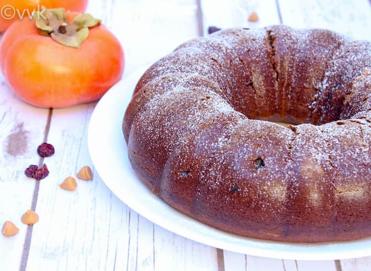 Eggless Persimmon Bundt Cake on a big white plate with more fruits on the side on a white wooden table