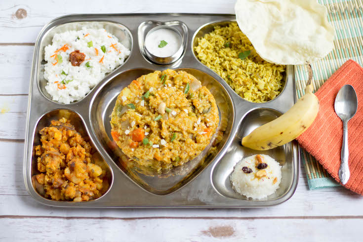 Saravana Bhavan Style Executive Thali with each dish titled on a wooden surface with a spoon next to the tray