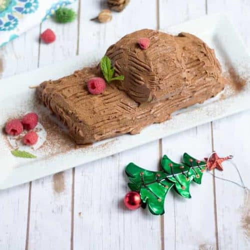 Christmas Special Eggless Yule Log Cake served on a white table with Christmas tree decorations around it