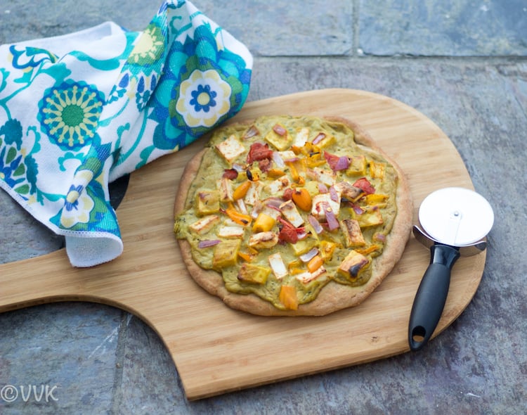 One big Shahi Paneer Pizza on board made of wood with a colorful towel and a rolling knife right next to it