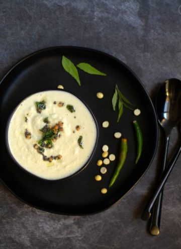 top angle shot of coconut chutney in a black bowl place on a black plate with green chilies and curry leaves on the side