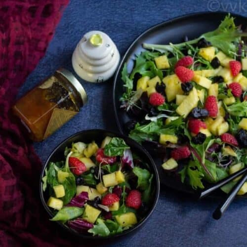 mixed greens and fruit salad in a black bowl and plate with honey on the side