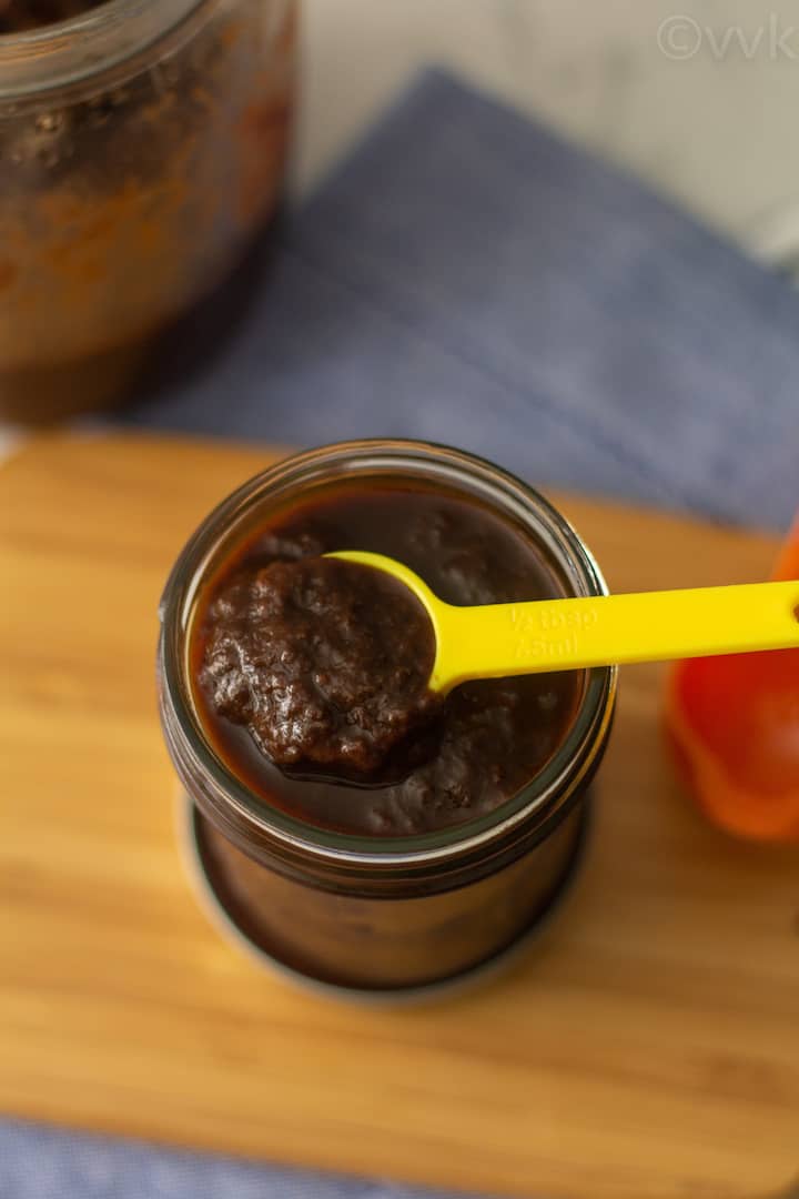 top angle shot of homemade tamarind paste in a glass jar placed on a wooden board with yellow spoon on top