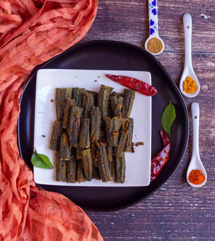 overhead shot of baked bhindi placed on white plate on top of black plate with assorted spices on the side and with dried red chilies