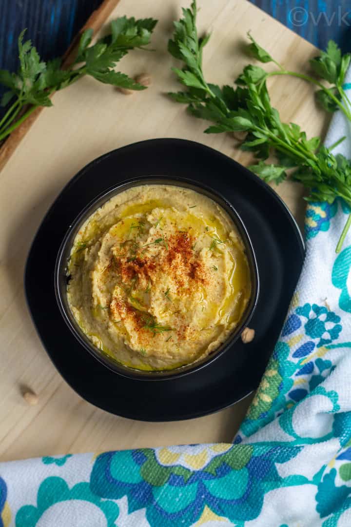 overhead shot of hummus placed in a black bowl on top of a black plate on a wooden board