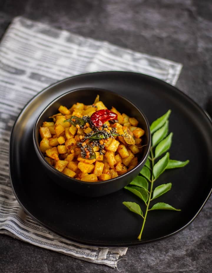 baked indian style potato curry served in a black bowl placed on a black plate with curry leaves on the side