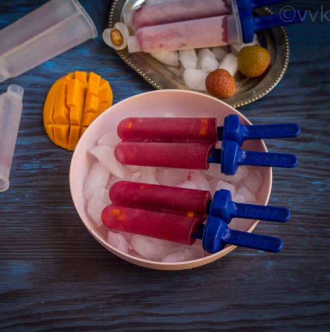 square image of black currant popsicle on a pink bowl with icecubes and chopped mango on the side