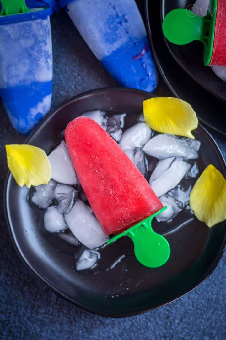 overhead shot of rose syrup rooh afza popsicle on a black plate with ice cubes and rose petals