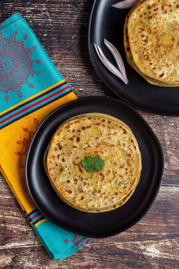 aloo paratha stacked up in black plates with onion wedges on one plate and cilantro on the other