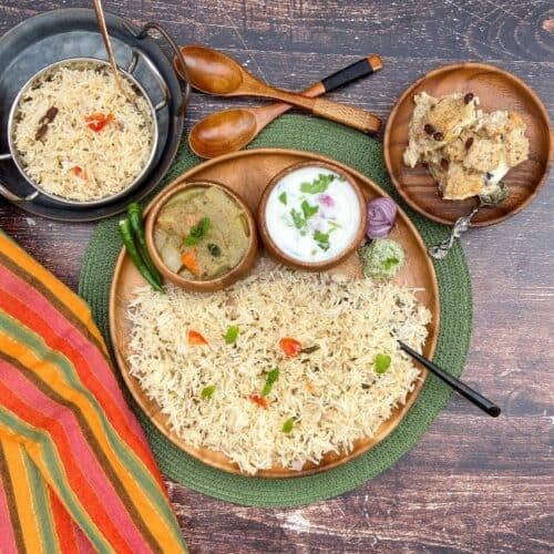 square image of overhead shot of coconut milk pulao platter with assorted sides served on a wooden plate