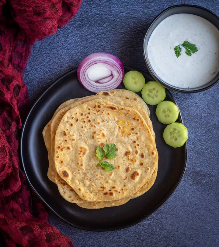 paneer paratha stacked in black plate with cut onions and cucumbers with raita on the side