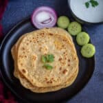 square image of paneer paratha with some raita, cut onions and cucumbers
