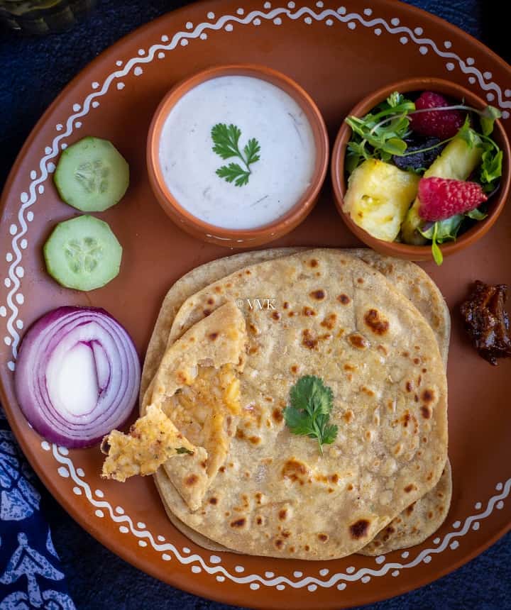close up shot of paneer paratha, raita, salad and pickle served on a terracotta plate