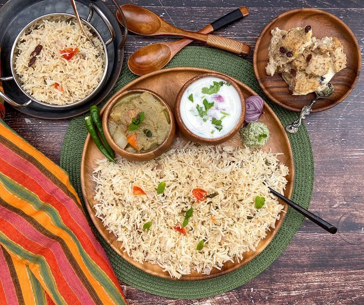 overhead shot of coconut milk pulao platter with assorted sides served on a wooden plate placed on a green mat