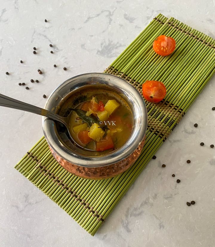 pineapple rasam in a traditional brassware placed on a green mat with tomatoes on the side