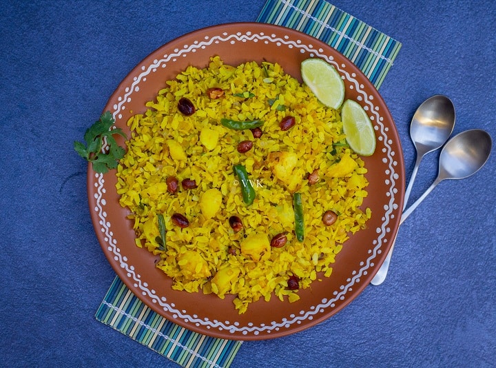 overhead shot of poha served in terracotta plate placed on a mat with spoons on the side and with some lemon wedges