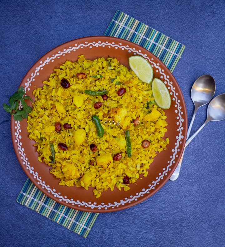 poha served in terracotta plate placed on a mat with spoons on the side and with some lemon wedges