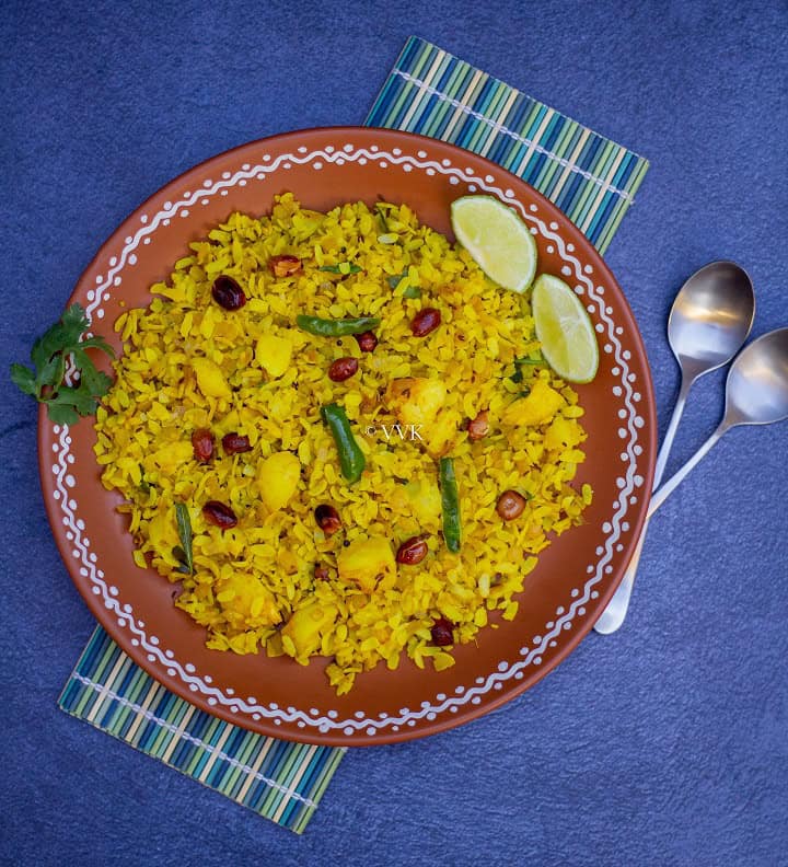 poha served in terracotta plate placed on a mat with spoons on the side and with some lemon wedges