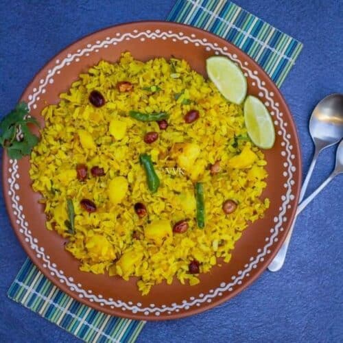 square image of poha served in terracotta plate placed on a mat with spoons on the side and with some lemon wedges