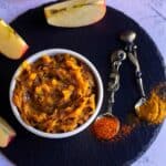 square image of overhead shot of apple pickle with apple slices on the side and with some red chili and turmeric powder sprinkled