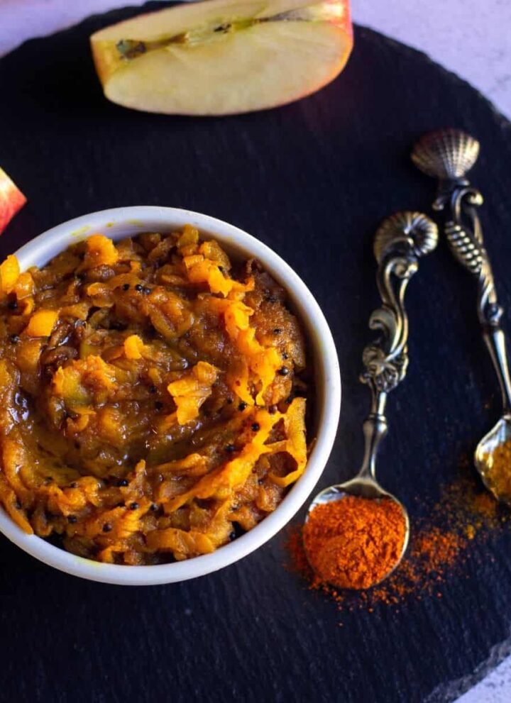square image of overhead shot of apple pickle with apple slices on the side and with some red chili and turmeric powder sprinkled