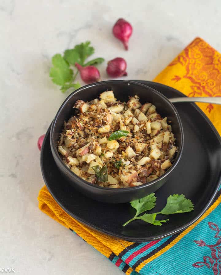 Chakkakuru thoran placed in black bowl on top of a plate with shallots on the top and cilantro in the side