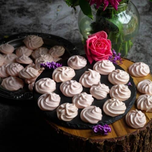 square image of vegan meringue cookies placed on a wooden stand with a flower vase behind