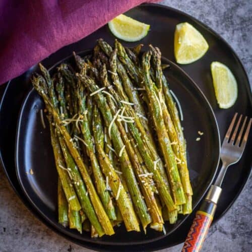 square image of baked asparagus served on a black plate with a fork on the side and some lemon wedges