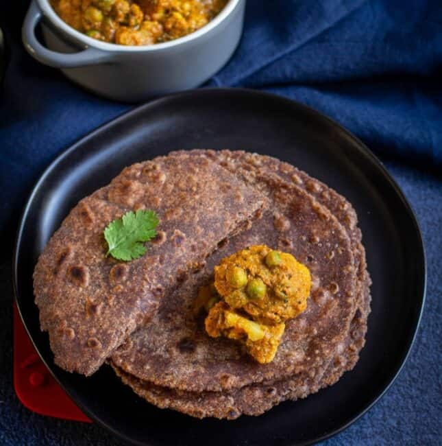 square image of ragi roti served on a black plate with cauliflower curry on top