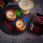 square image of thandai served in kulhad cups and glass cup placed on a black board with colors