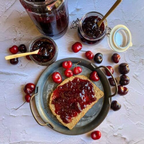 overhead shot of bread smeared with cherry jam with jam bottles and fresh cherries on the side