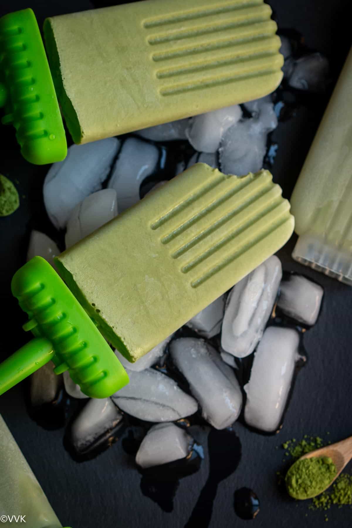 two green tea matcha popsicles  with one bitten placed on ice cubes on a black slate board