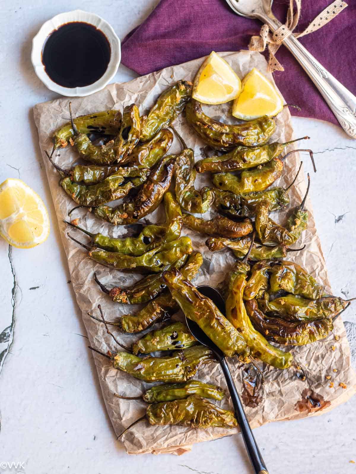 roasted shishito peppers served in a slate board wrapped with parchment paper with lemon wedges and soy sauce on the side