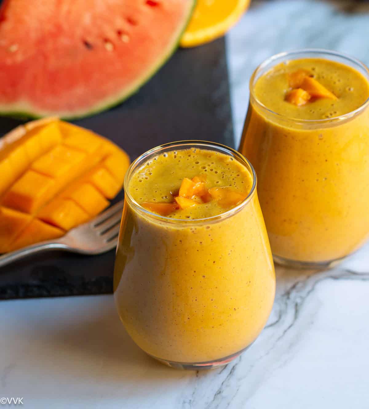 close up shot of mango smoothie topped with mangoes with some fruits on the side placed on a black slate board