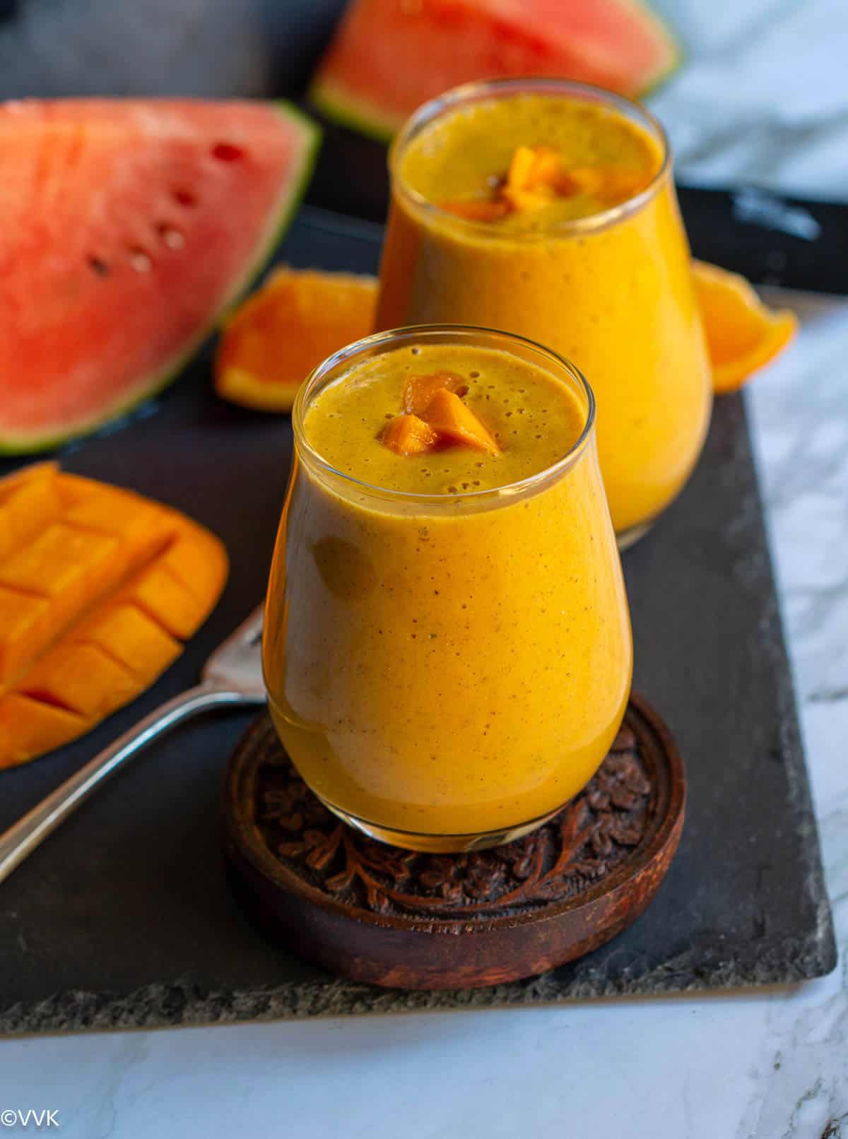 mango breakfast smoothie served in a glass topped with freshly cut mangoes placed on wooden coaster placed on a slate board