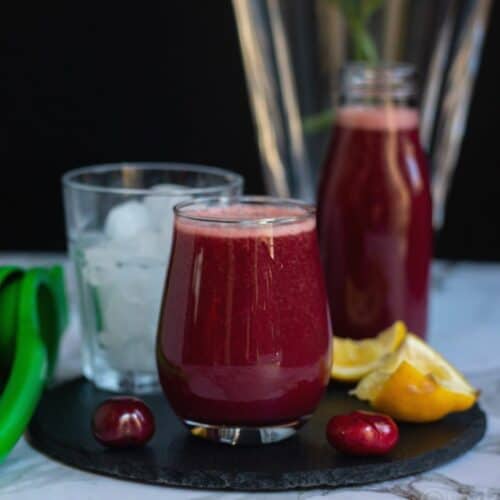 cherry lemonade served in a glass with the syrup behind and a glass filling with ice.