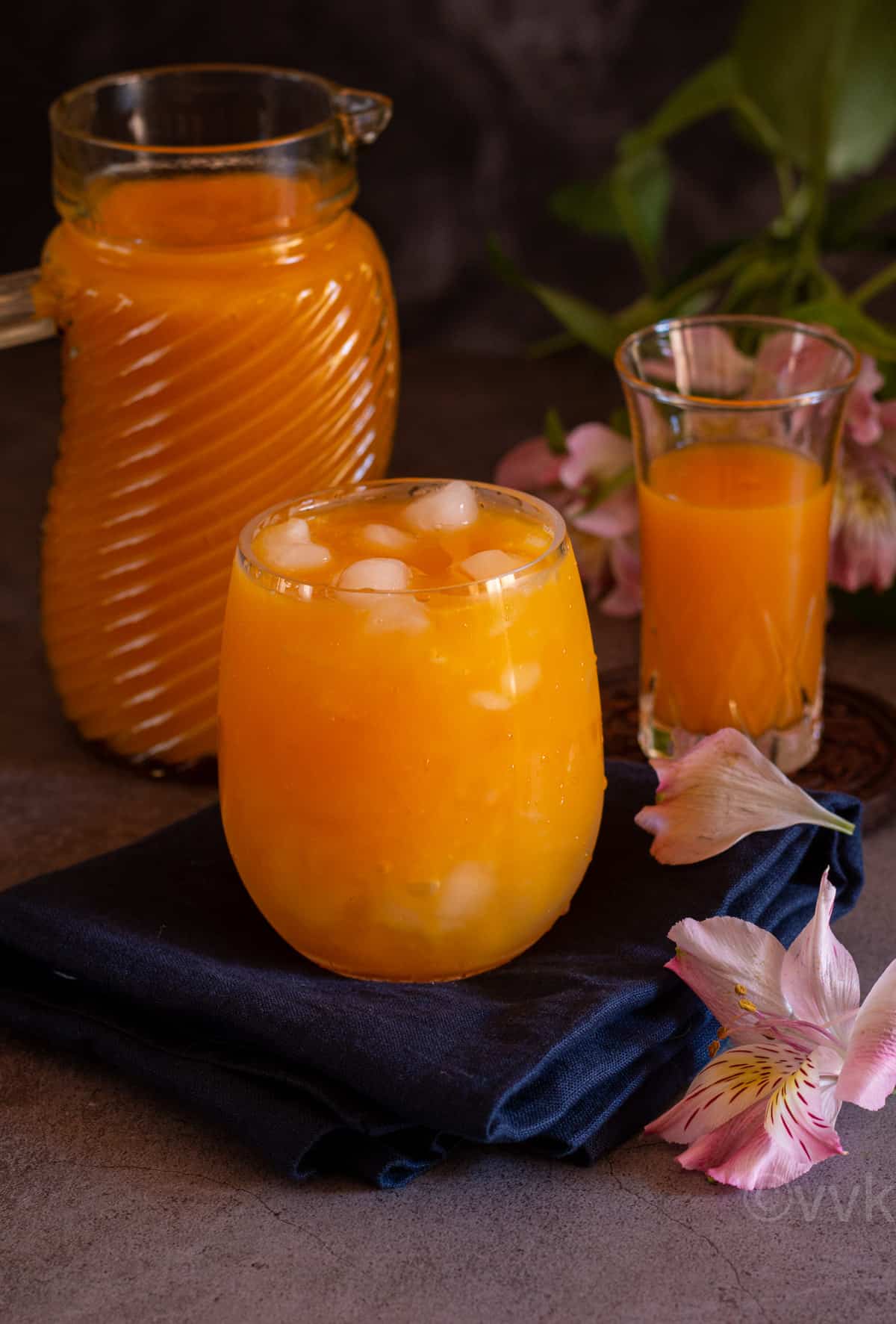fresh mango nectar or mango fruity served in tall glass with ice cubes with flowers on the side and mango puree behind