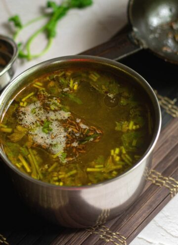 square image of rasam served in stainless vessel placed on a mat with a tempering spoon on the side
