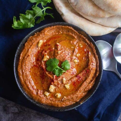 close up shot of muhammara served in bowl with pita bread on the side placed on a blue towel