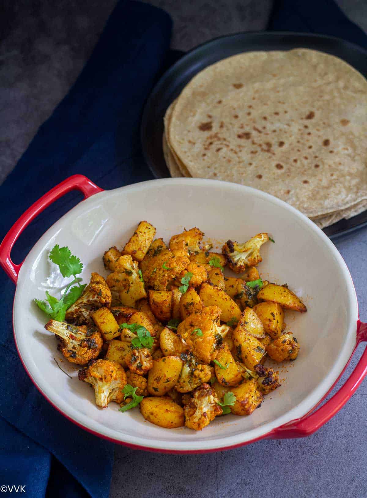 aloo gobi served in red color stone ware wrapped with blue towel with roti on the side