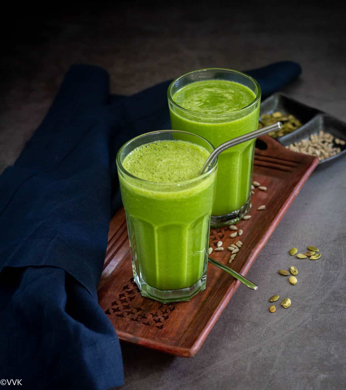 spinach smoothie with apple and banana served in two glasses placed on wooden tray with seeds sprinkled