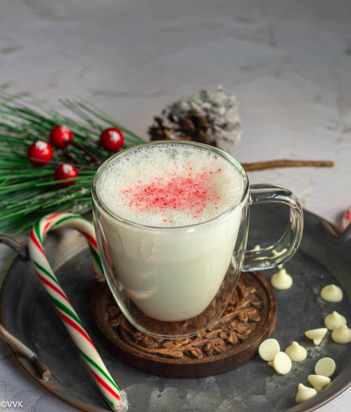 white hot chocolate served in a mug placed on a wooden coaster with some candy canes and choco chips on the side