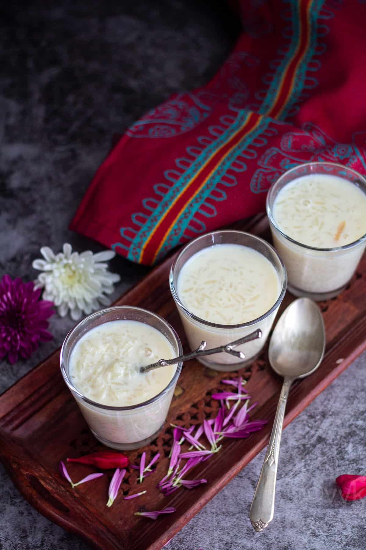 arisi pal payasam served in three glass tumblers with flowers on the side on a wooden board