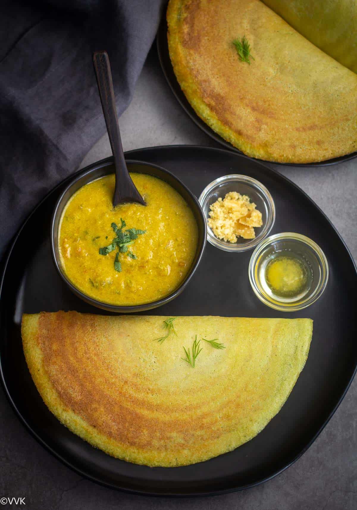 overhead shot of shepucha dosa served with kurma, jaggery and ghee on a black plate 