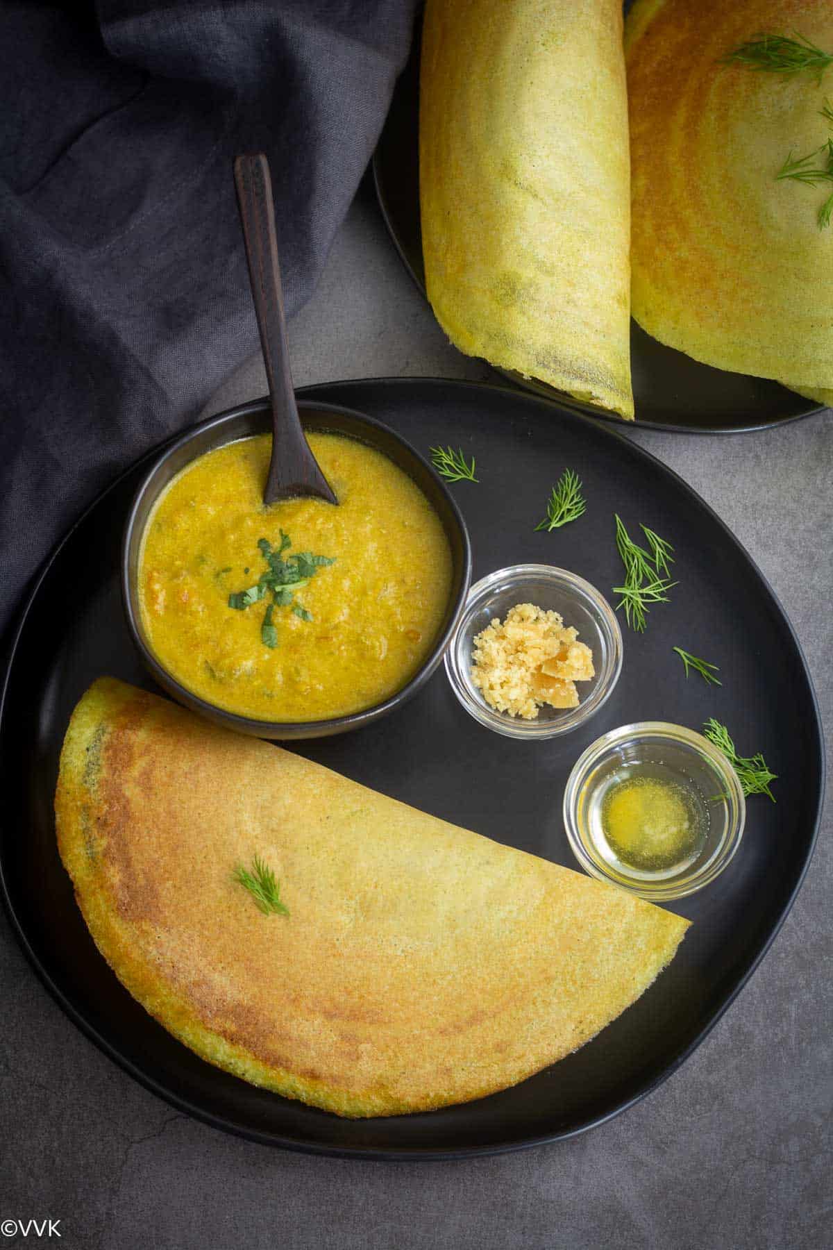 dill weed dosa served with kurma, jaggery and ghee in a black plate with more dosa on the side